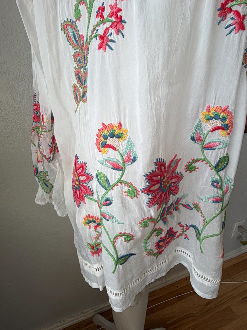 May include: White tunic with colorful floral embroidery. The embroidery features red, pink, yellow, and green flowers and leaves. The tunic has a loose fit and a scalloped hemline. The fabric appears to be lightweight and slightly sheer.