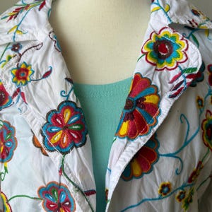 Puede incluir: Chaqueta blanca con bordado floral colorido. La chaqueta tiene cuello y cierre de botones. El bordado incluye flores y enredaderas rojas, amarillas, azules y verdes. Se ve una camiseta azul claro debajo.