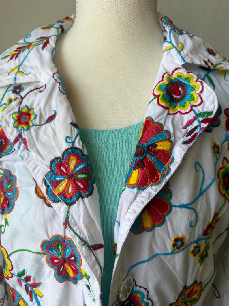 Puede incluir: Chaqueta blanca con bordado floral colorido. La chaqueta tiene cuello y cierre de botones. El bordado incluye flores y enredaderas rojas, amarillas, azules y verdes. Se ve una camiseta azul claro debajo.