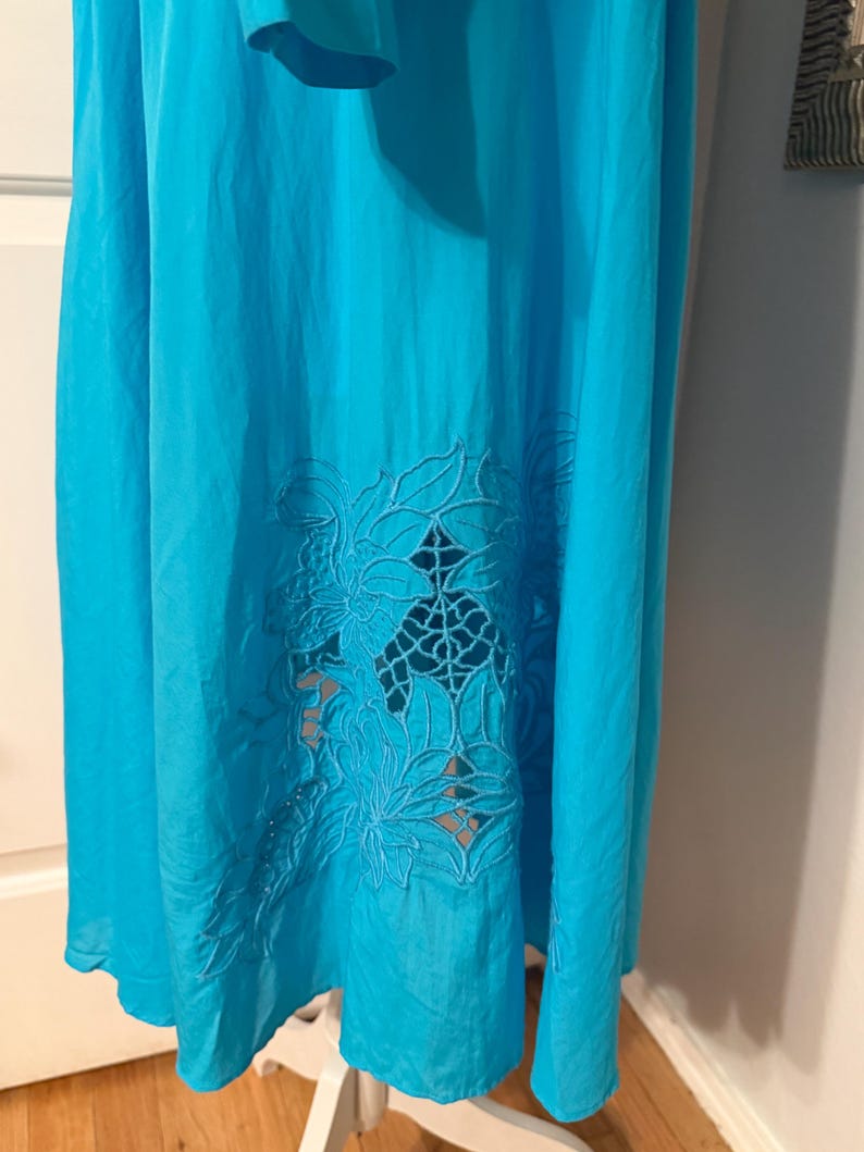 Puede incluir: Un vestido turquesa vibrante con un dise&ntilde;o floral recortado cerca del dobladillo. El vestido parece estar hecho de una tela ligera, adecuada para climas c&aacute;lidos. El patr&oacute;n recortado a&ntilde;ade un elemento decorativo a la prenda.