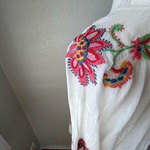 May include: White garment with vibrant floral embroidery in red, pink, yellow, and blue. The embroidered design features large flowers and stems, creating a colorful pattern. The fabric appears to be lightweight and textured.
