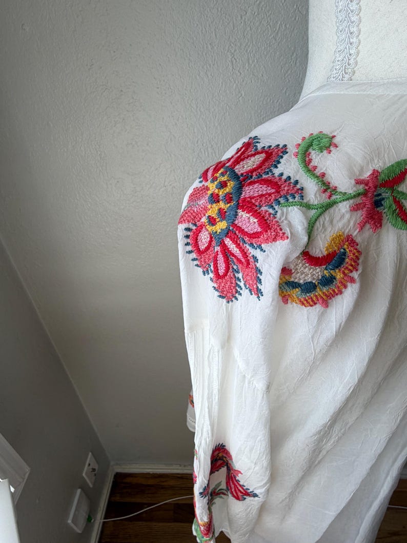 May include: White garment with vibrant floral embroidery in red, pink, yellow, and blue. The embroidered design features large flowers and stems, creating a colorful pattern. The fabric appears to be lightweight and textured.