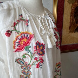 May include: White tunic top with colorful floral embroidery. The embroidery features red, pink, yellow, blue, and green flowers and leaves. The top has a drawstring neckline with white tassels.