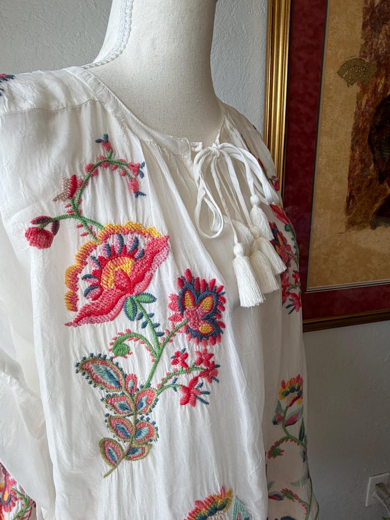 May include: White tunic top with colorful floral embroidery. The embroidery features red, pink, yellow, blue, and green flowers and leaves. The top has a drawstring neckline with white tassels.
