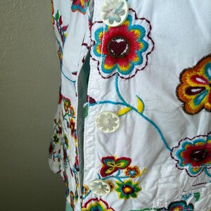 Puede incluir: Chaqueta blanca bordada con dise&ntilde;os florales coloridos. La chaqueta presenta una parte delantera abotonada con botones en forma de flor. El bordado incluye patrones florales rojos, amarillos, azules y verdes, creando un aspecto vibrante y art&iacute;stico.