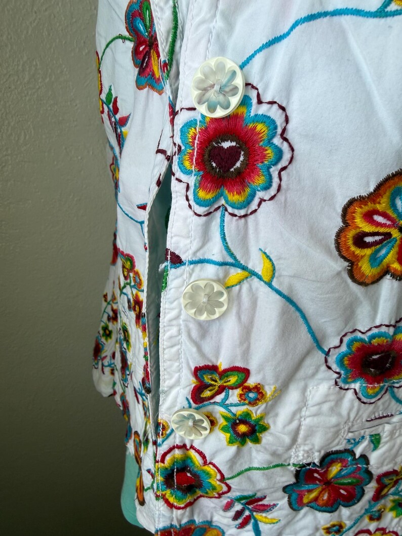 Puede incluir: Chaqueta blanca bordada con dise&ntilde;os florales coloridos. La chaqueta presenta una parte delantera abotonada con botones en forma de flor. El bordado incluye patrones florales rojos, amarillos, azules y verdes, creando un aspecto vibrante y art&iacute;stico.