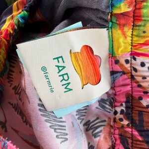 Puede incluir: Primer plano de tela colorida con estampado floral y abstracto. La tela tiene una etiqueta con la palabra "FARM" en verde, un logotipo colorido y el identificador "@farmrio". La tela es una mezcla de rosa, amarillo, azul y negro.