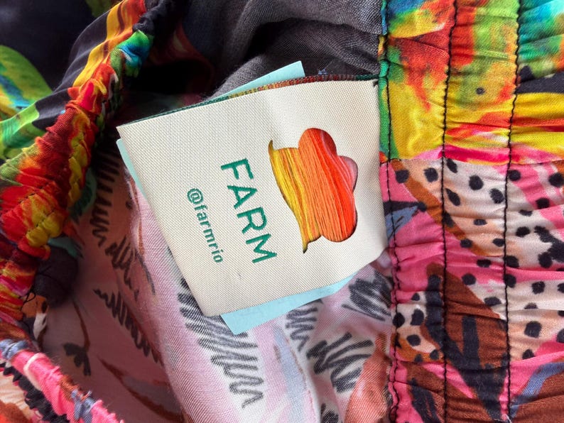 Puede incluir: Primer plano de tela colorida con estampado floral y abstracto. La tela tiene una etiqueta con la palabra "FARM" en verde, un logotipo colorido y el identificador "@farmrio". La tela es una mezcla de rosa, amarillo, azul y negro.