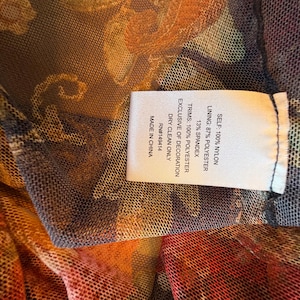 May include: Close-up of a sheer fabric with a floral pattern in shades of orange, red, and brown. A white tag with black text details the fabric composition: 100% nylon, 87% polyester, and 13% spandex. The tag also indicates dry clean only.