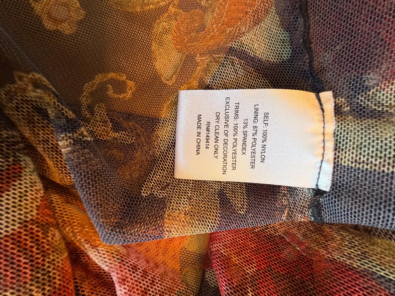 May include: Close-up of a sheer fabric with a floral pattern in shades of orange, red, and brown. A white tag with black text details the fabric composition: 100% nylon, 87% polyester, and 13% spandex. The tag also indicates dry clean only.