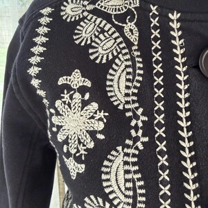 K&ouml;nnte beinhalten: Schwarze Jacke mit aufwendigen wei&szlig;en Stickereien mit Blumen- und Paisley-Mustern. Die Jacke hat einen hohen Kragen und Knopfverschl&uuml;sse. Die Stickerei bedeckt Schultern, Vorderseite und &Auml;rmel.