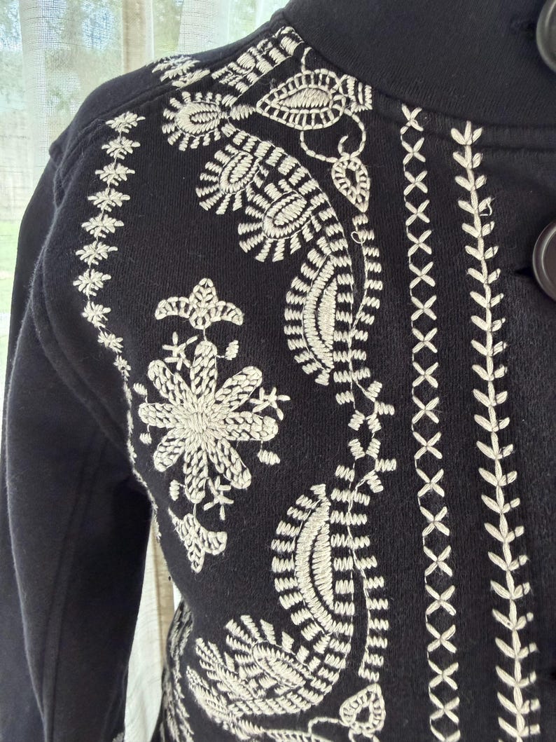 K&ouml;nnte beinhalten: Schwarze Jacke mit aufwendigen wei&szlig;en Stickereien mit Blumen- und Paisley-Mustern. Die Jacke hat einen hohen Kragen und Knopfverschl&uuml;sse. Die Stickerei bedeckt Schultern, Vorderseite und &Auml;rmel.