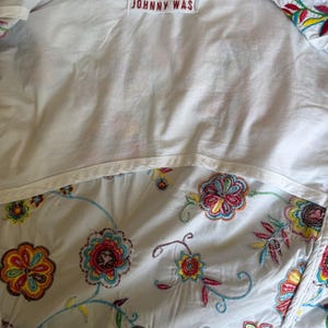 Puede incluir: Camisa blanca con bordados florales coloridos. El interior de la camisa es blanco con una etiqueta "JOHNNY WAS". El bordado floral incluye hilo rojo, amarillo, azul y verde.