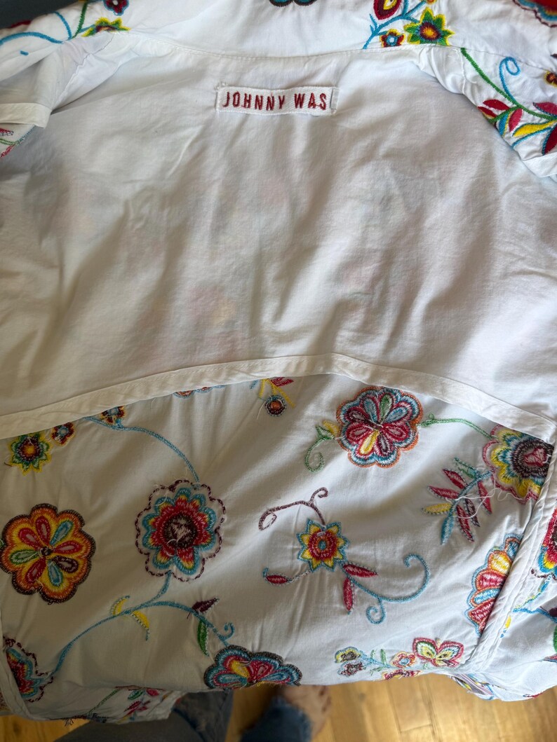Puede incluir: Camisa blanca con bordados florales coloridos. El interior de la camisa es blanco con una etiqueta "JOHNNY WAS". El bordado floral incluye hilo rojo, amarillo, azul y verde.