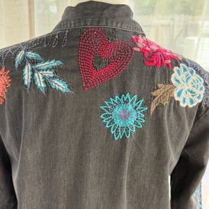 以下が含まれることがあります： 背中にカラフルな花の刺繍が施された濃いグレーのデニムジャケット。刺繍には赤いハート、青とターコイズの葉、ピンク、オレンジ、青の色合いの花が含まれています。