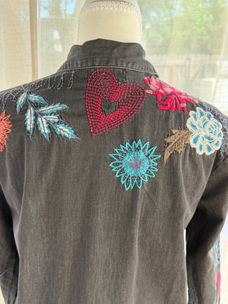 以下が含まれることがあります： 背中にカラフルな花の刺繍が施された濃いグレーのデニムジャケット。刺繍には赤いハート、青とターコイズの葉、ピンク、オレンジ、青の色合いの花が含まれています。