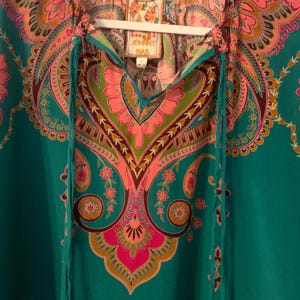 May include: A teal tunic top with a colorful paisley pattern in pink, green, and brown. The top has a V-neckline with teal drawstrings and a tag that reads "JOHNNY WAS". The tunic is hanging on a white hanger.