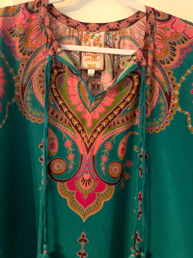 May include: A teal tunic top with a colorful paisley pattern in pink, green, and brown. The top has a V-neckline with teal drawstrings and a tag that reads "JOHNNY WAS". The tunic is hanging on a white hanger.