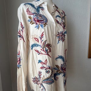 May include: Cream-colored button-down shirt with long sleeves, featuring intricate floral embroidery in shades of blue, red, and brown. The embroidery is detailed and covers the front and sleeves of the shirt. The shirt is displayed on a mannequin.