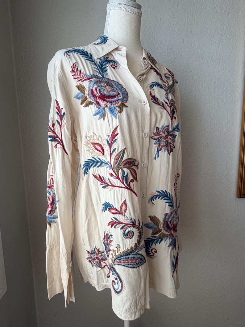 May include: Cream-colored button-down shirt with long sleeves, featuring intricate floral embroidery in shades of blue, red, and brown. The embroidery is detailed and covers the front and sleeves of the shirt. The shirt is displayed on a mannequin.
