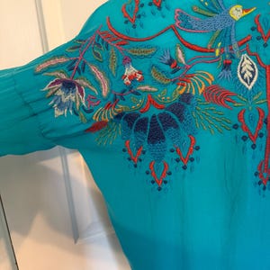 May include: Teal-colored blouse with intricate embroidery. The embroidery features a bird, flowers, and foliage in shades of blue, red, and green. The design is on the back and sleeve of the blouse. The blouse has long sleeves and a button-down front.