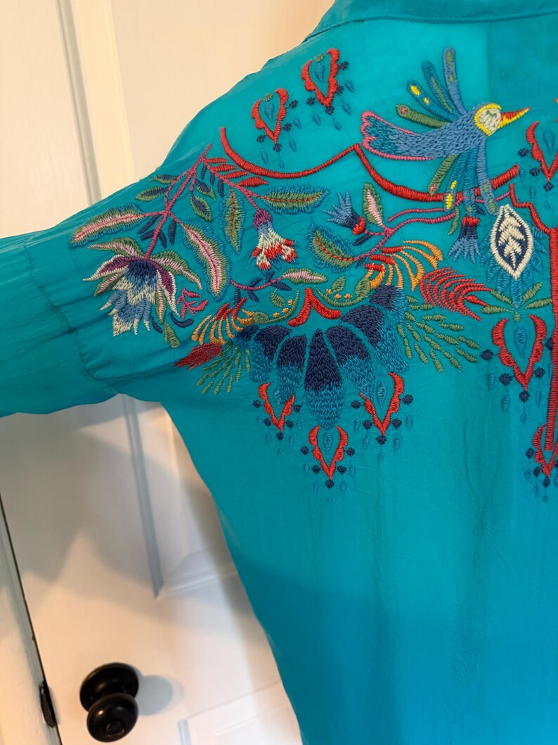 May include: Teal-colored blouse with intricate embroidery. The embroidery features a bird, flowers, and foliage in shades of blue, red, and green. The design is on the back and sleeve of the blouse. The blouse has long sleeves and a button-down front.