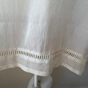 May include: Off-white fabric with a textured appearance. The bottom edge features a decorative cut-out design. The fabric is draped over a mannequin, suggesting a garment.