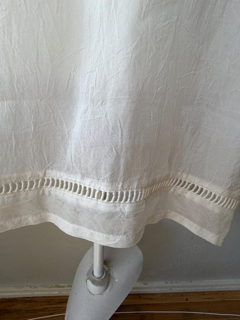 May include: Off-white fabric with a textured appearance. The bottom edge features a decorative cut-out design. The fabric is draped over a mannequin, suggesting a garment.