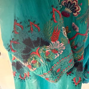 May include: Teal-colored embroidered tunic with floral and leaf patterns in red, green, and blue. The embroidery is concentrated on the sleeves and upper chest area, creating a vibrant contrast against the solid teal fabric.
