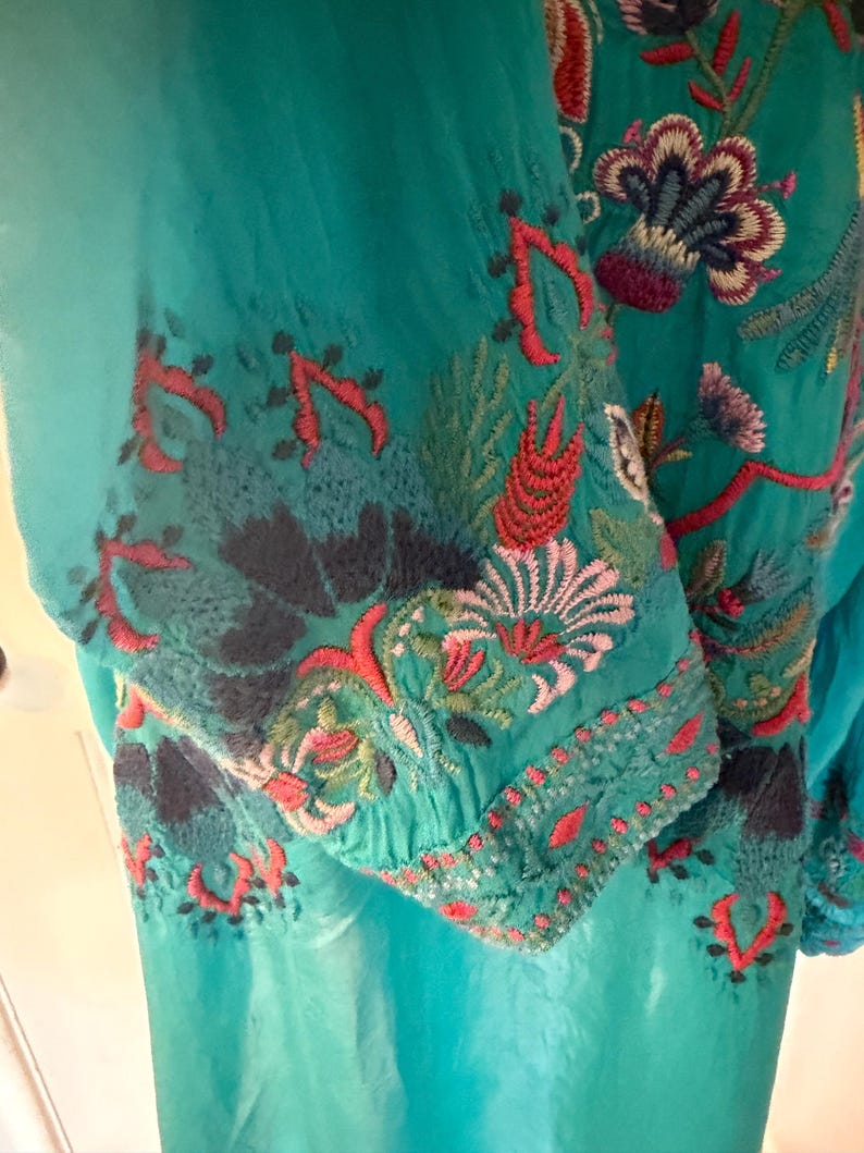 May include: Teal-colored embroidered tunic with floral and leaf patterns in red, green, and blue. The embroidery is concentrated on the sleeves and upper chest area, creating a vibrant contrast against the solid teal fabric.