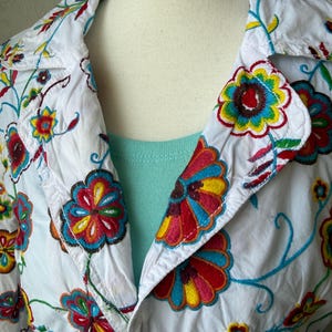 Puede incluir: Chaqueta blanca con bordado floral colorido. La chaqueta tiene cuello y cierre de bot&oacute;n. El bordado incluye flores y enredaderas rojas, amarillas, azules y verdes.