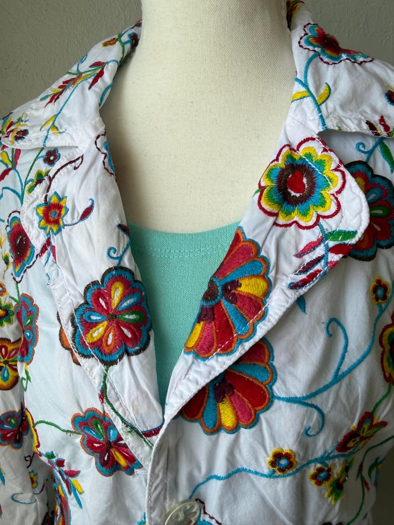 Puede incluir: Chaqueta blanca con bordado floral colorido. La chaqueta tiene cuello y cierre de bot&oacute;n. El bordado incluye flores y enredaderas rojas, amarillas, azules y verdes.