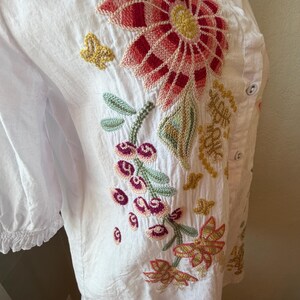 May include: White button-down shirt with colorful floral embroidery. The embroidery features a large red and pink flower, green leaves, and smaller flowers in shades of pink, yellow, and orange. The shirt has a ruffled sleeve detail.