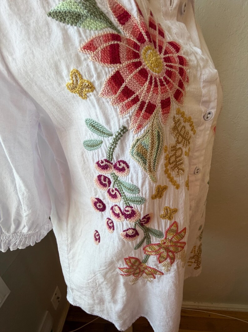 May include: White button-down shirt with colorful floral embroidery. The embroidery features a large red and pink flower, green leaves, and smaller flowers in shades of pink, yellow, and orange. The shirt has a ruffled sleeve detail.