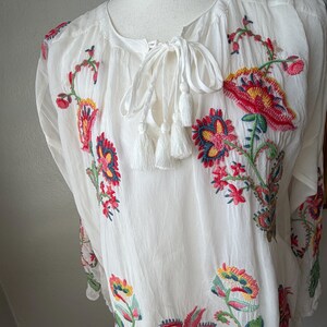 May include: White tunic top with colorful floral embroidery. The embroidered flowers feature red, yellow, and blue details. The top has a tie-neck closure with white tassels.