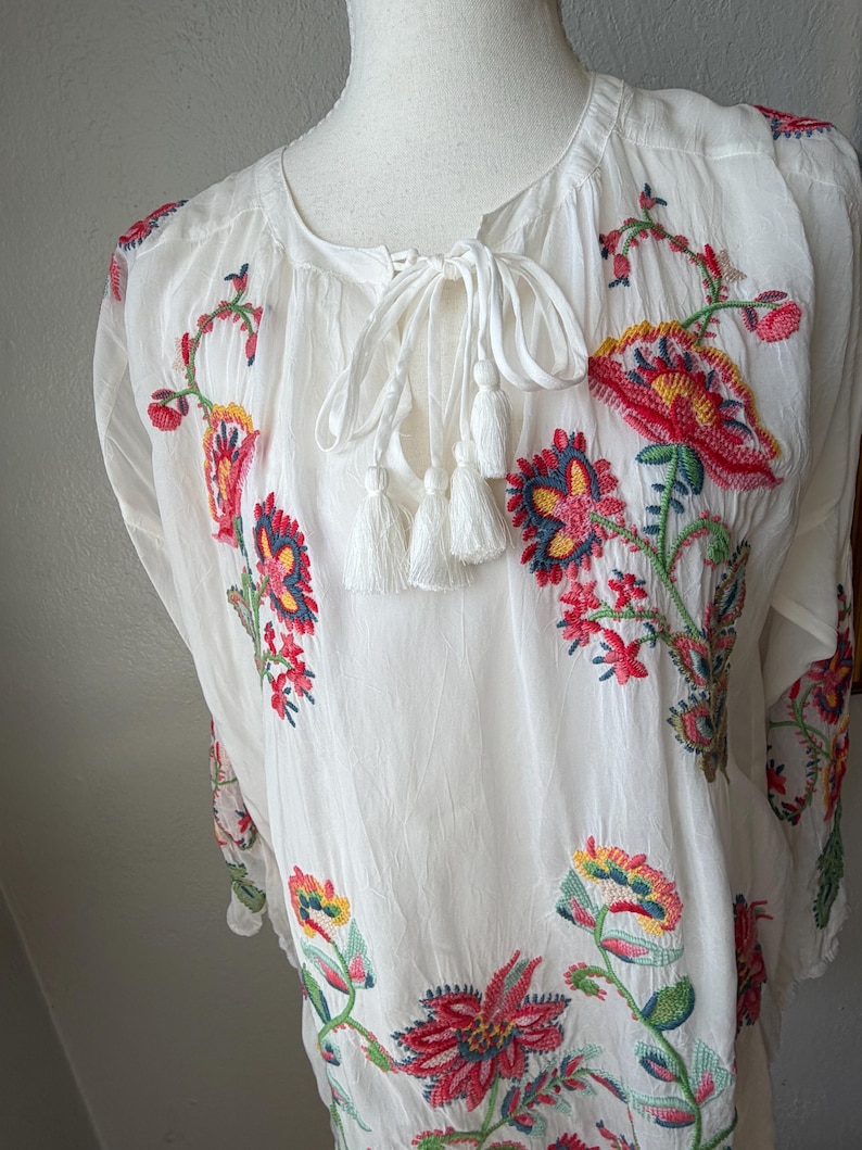 May include: White tunic top with colorful floral embroidery. The embroidered flowers feature red, yellow, and blue details. The top has a tie-neck closure with white tassels.