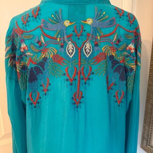 May include: A turquoise long-sleeve shirt with intricate embroidery on the back. The embroidery features colorful birds, leaves, and floral patterns in shades of red, blue, yellow, and green. The shirt has a collared neckline.