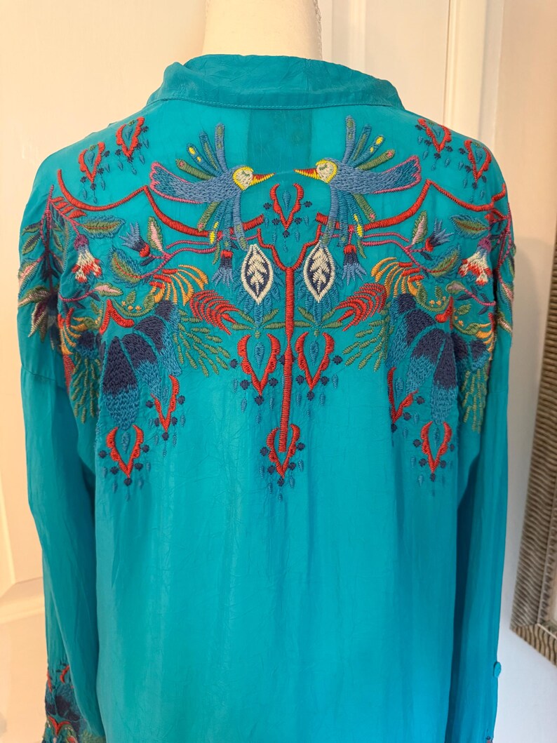 May include: A turquoise long-sleeve shirt with intricate embroidery on the back. The embroidery features colorful birds, leaves, and floral patterns in shades of red, blue, yellow, and green. The shirt has a collared neckline.