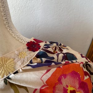 May include: Close-up of a floral print garment. The fabric features large, vibrant flowers in shades of orange, pink, and red against a cream background. The neckline is trimmed with embroidered details.