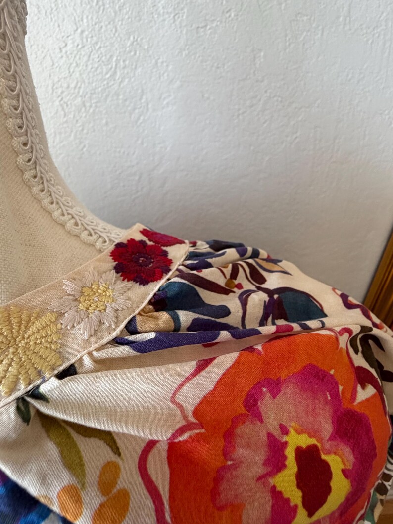 May include: Close-up of a floral print garment. The fabric features large, vibrant flowers in shades of orange, pink, and red against a cream background. The neckline is trimmed with embroidered details.