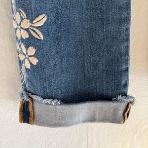 Op de afbeelding: Close-up van een blauwe denim jeans met een bloemenborduurontwerp aan de zijkant. De jeans heeft een opgerolde zoom met een gerafelde rand en een contrasterend goud en marineblauw detail aan de binnenkant van de manchet.