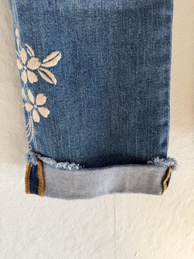 Op de afbeelding: Close-up van een blauwe denim jeans met een bloemenborduurontwerp aan de zijkant. De jeans heeft een opgerolde zoom met een gerafelde rand en een contrasterend goud en marineblauw detail aan de binnenkant van de manchet.
