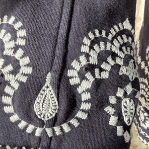 K&ouml;nnte beinhalten: Nahaufnahme eines marineblauen Kleidungsst&uuml;cks mit detaillierten wei&szlig;en Stickereien. Das Design umfasst florale und geometrische Muster, wie z. B. verbundene Schlaufen und Kreuzstichakzente. Der Stoff hat ein strukturiertes Aussehen.