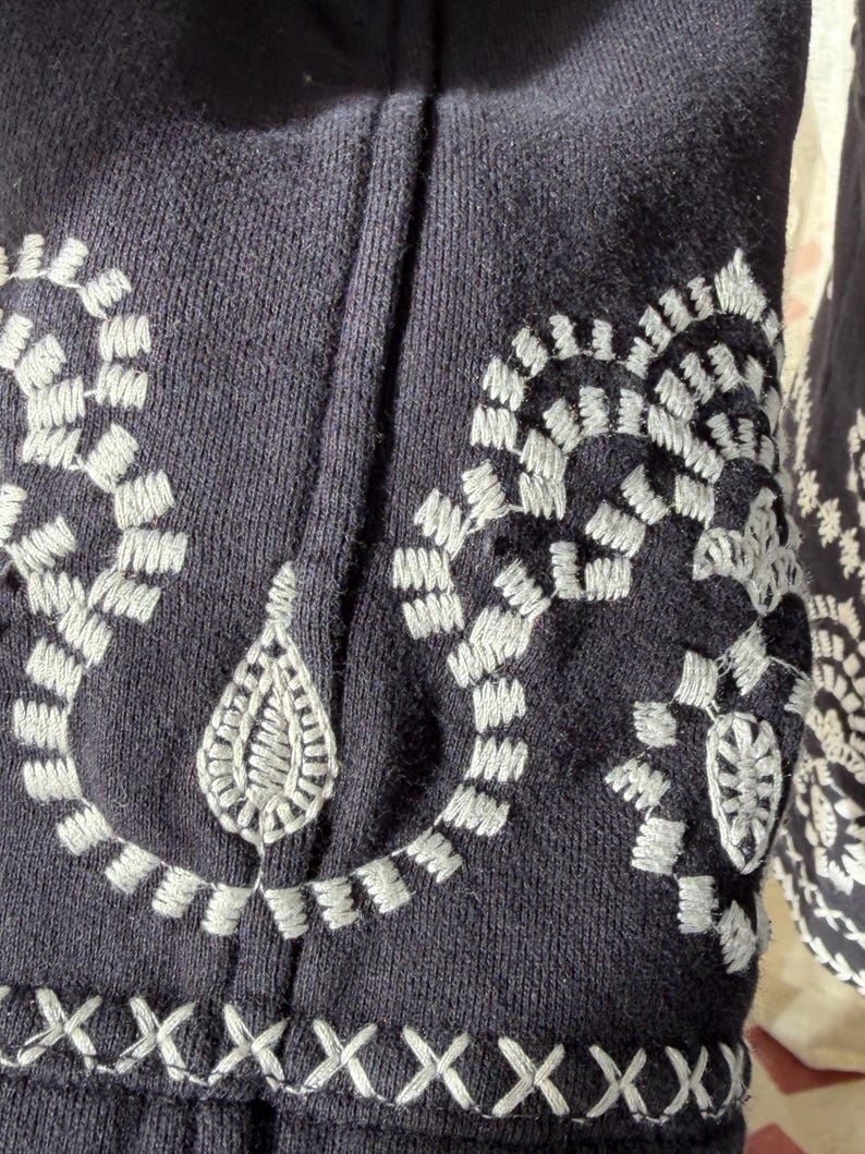 K&ouml;nnte beinhalten: Nahaufnahme eines marineblauen Kleidungsst&uuml;cks mit detaillierten wei&szlig;en Stickereien. Das Design umfasst florale und geometrische Muster, wie z. B. verbundene Schlaufen und Kreuzstichakzente. Der Stoff hat ein strukturiertes Aussehen.