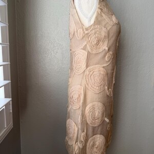 May include: A sleeveless, beige dress with a V-neck and a unique floral design. The dress features multiple, large, circular rosettes, adding texture and visual interest. The fabric appears lightweight and flowing, suitable for warmer weather.