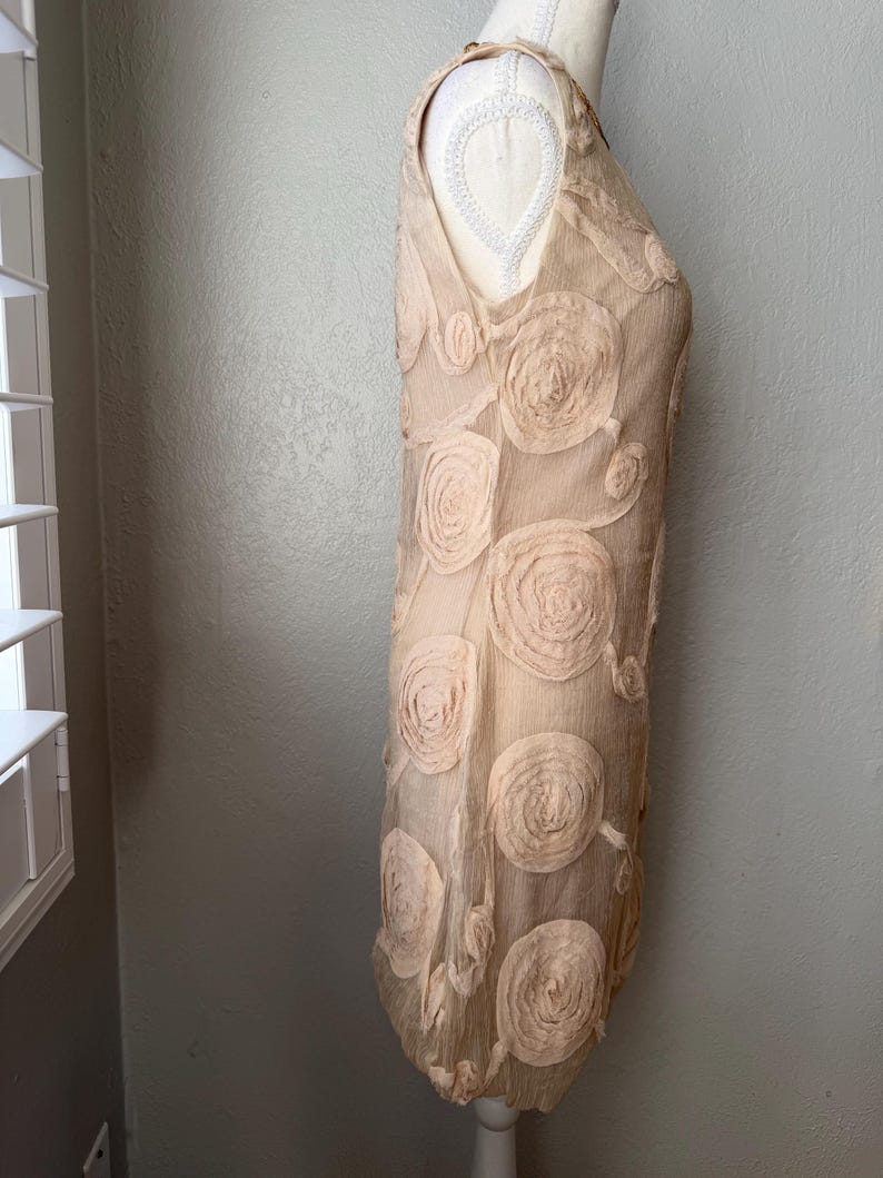 May include: A sleeveless, beige dress with a V-neck and a unique floral design. The dress features multiple, large, circular rosettes, adding texture and visual interest. The fabric appears lightweight and flowing, suitable for warmer weather.