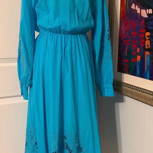 Puede incluir: Un vestido largo turquesa con mangas largas y cintura ce&ntilde;ida. Las mangas tienen calados bordados y la falda presenta calados florales cerca del dobladillo. El vestido se exhibe en un maniqu&iacute;.