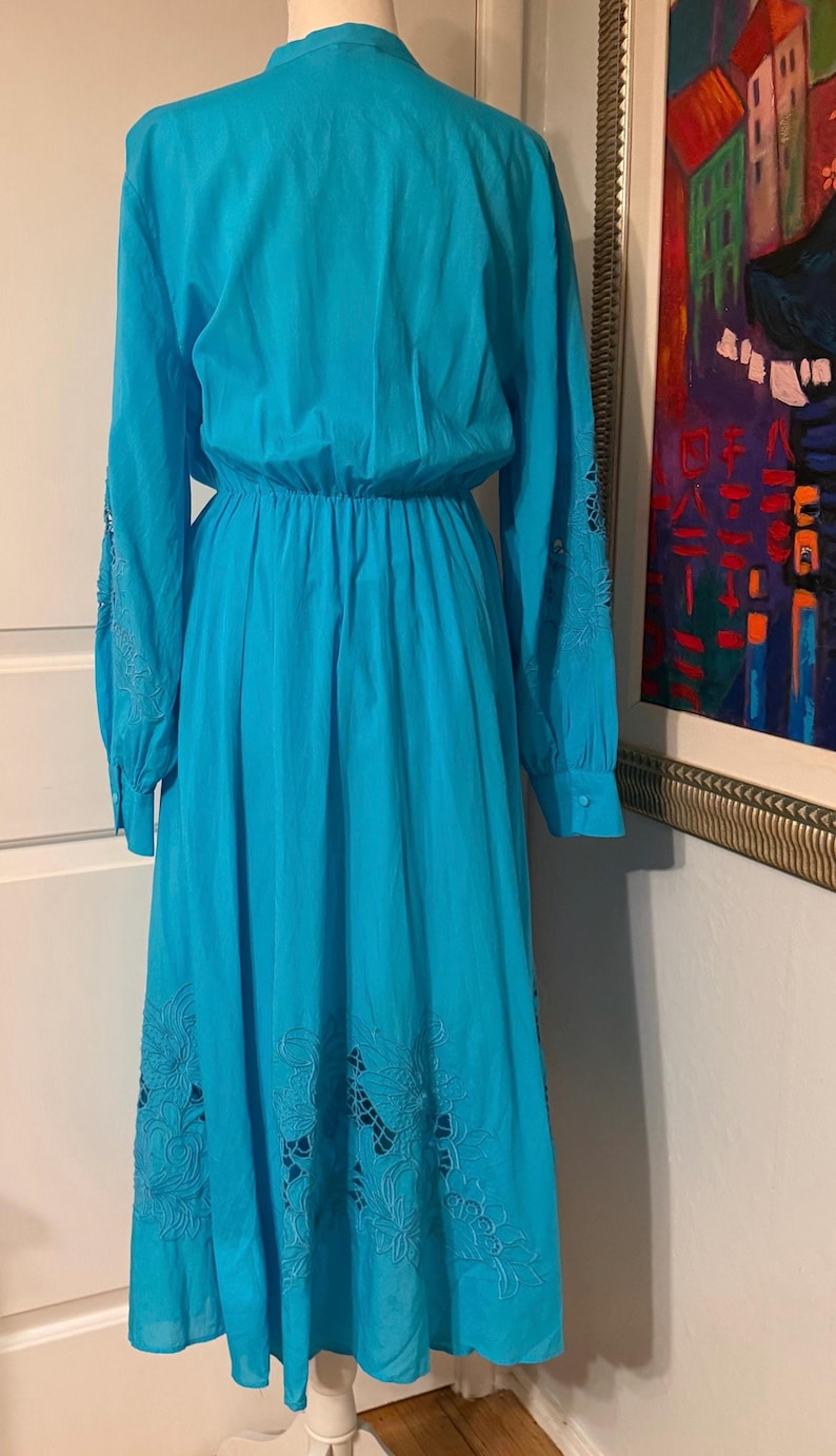 Puede incluir: Un vestido largo turquesa con mangas largas y cintura ce&ntilde;ida. Las mangas tienen calados bordados y la falda presenta calados florales cerca del dobladillo. El vestido se exhibe en un maniqu&iacute;.