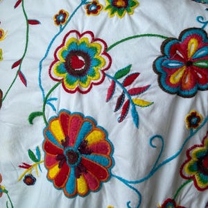 Puede incluir: Primer plano de tela blanca adornada con bordados florales coloridos. El bordado presenta grandes flores multicolores en tonos rojos, amarillos, azules y verdes, con delicadas vides y hojas.