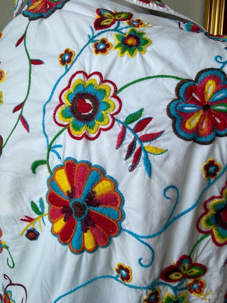 Puede incluir: Primer plano de tela blanca adornada con bordados florales coloridos. El bordado presenta grandes flores multicolores en tonos rojos, amarillos, azules y verdes, con delicadas vides y hojas.